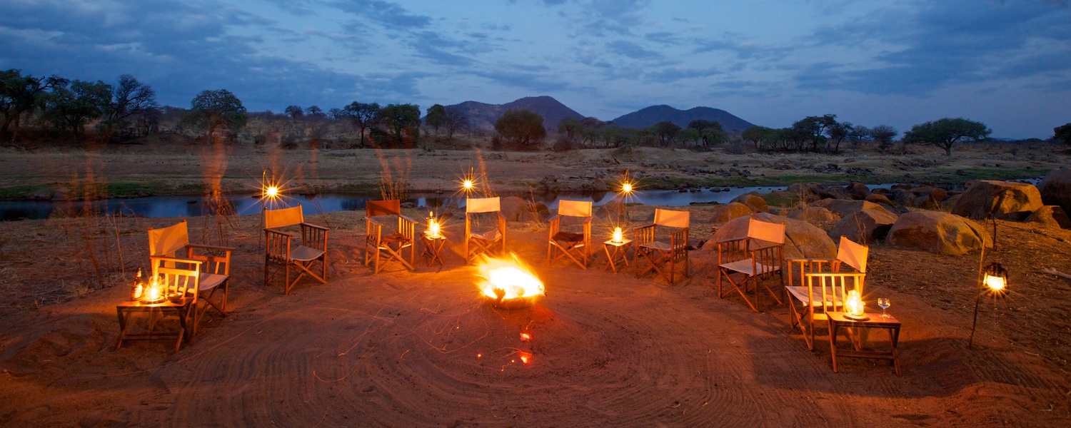 Ruaha River Lodge camp fire at dust. The camp fire is alight and safari chairs are surrounding it. This camp fire is positioned next to The Great Ruaha River in Ruaha National Park